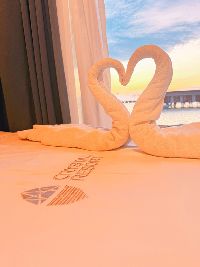 A cozy bed with towel art and resort logo next to glass doors revealing a vibrant sunset view over the Semporna islands at Crystal Resort